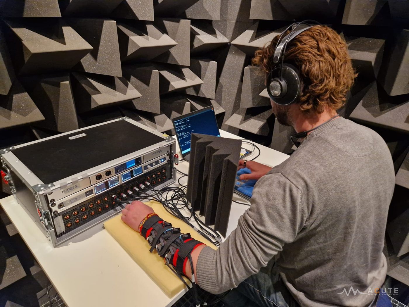 A participant wearing a vibrotactile sleeve with one hand resting on a cushion, headphones for white noise, and using a keypad to reproduce Braille-like vibrotactile patterns generated by the sleeve.